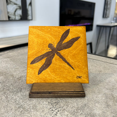 A laser engraved dragonfly on an amber stain baltic birch art tile with an earth colour baltic birch display stand