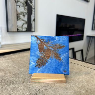 Laser engraved pine bough on blue stain baltic birch tile displayed on a natural baltic birch stand